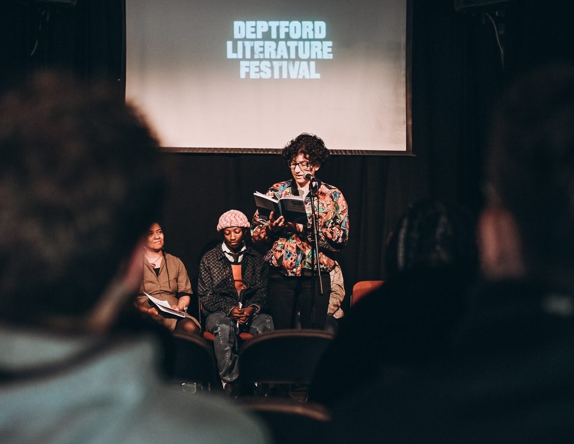 Giselle Cory reading her work at a microphone on stage at the Deptford Literature Festival, with other writers seated behind her and the festival name on a projector screen above.