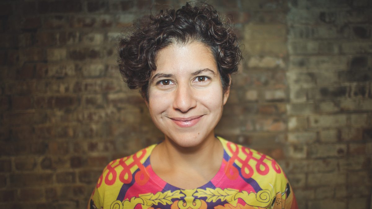 Portrait of Giselle Cory smiling, wearing a brightly patterned top against a brick wall.
